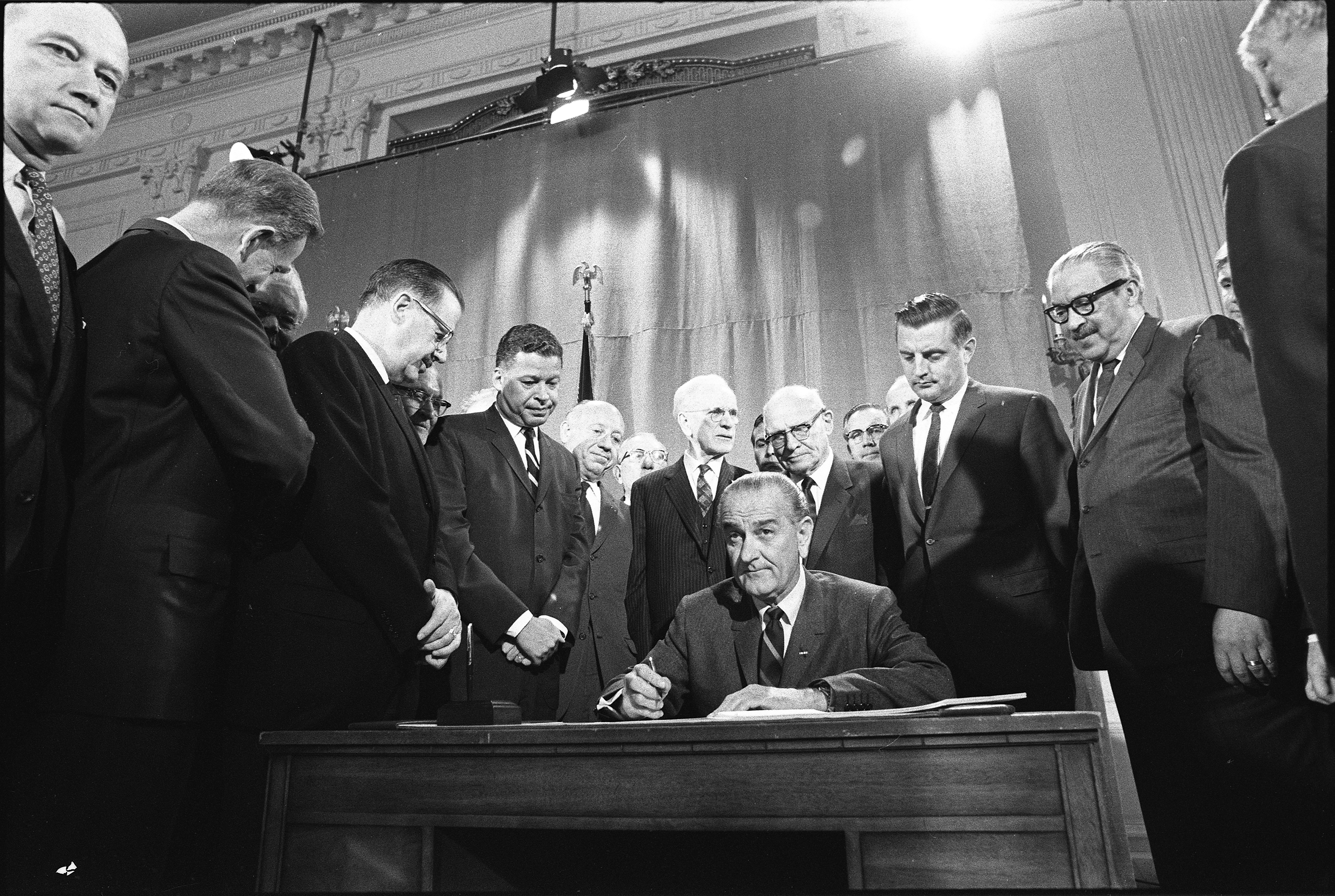 President Johnson, seated at desk, looks up from signing a piece of paper while a crowd of men in suits observe. 